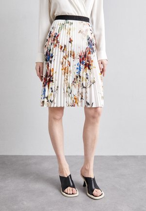Pleated white skirt with multi-colored floral print, worn with a white long-sleeve top and black open-toe heeled sandals.