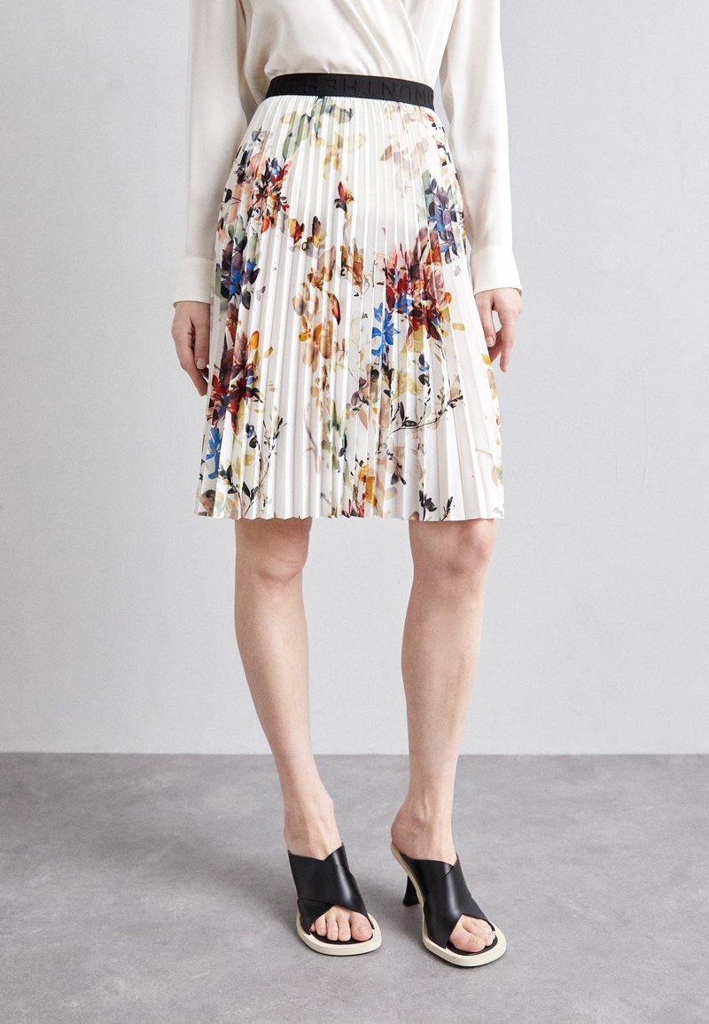 Jupe blanche plissée avec imprimé floral multicolore, portée avec un top blanc à manches longues et des sandales à talons noires à bouts ouverts.