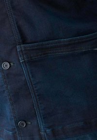 Donkerdenimjack met knoopsluiting, enkele borstzak, contrasterende stiksels en een lichtgetextureerde afwerking.