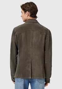 Giacca in suede marrone con una vestibilità su misura, caratterizzata da una fessura sul retro, polsini con bottone e una texture morbida, abbinata a jeans azzurri chiari.