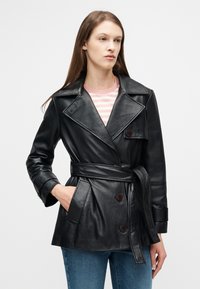 Γυναίκα που φοράει μαύρο δερμάτινο μπουφάν με ζώνη, ριγέ ροζ και λευκό πουκάμισο και μπλε τζιν, με το ένα χέρι στην τσέπη του μπουφάν.