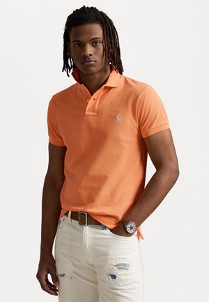 Oranje polo shirt met een kraag, twee knopen en een klein blauw logo. Draagt het met versleten witte spijkerbroeken en een bruine riem. Horloge om de pols.