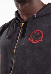 Zwarte hoodie met rits en gouden ritsen, voorzien van een verstelbaar trekkoord. Heeft een gestructureerde stof en een cirkelvormig rood en zwart logo op de borst.