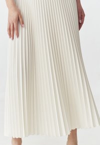 Gonna midi plissettata di colore crema. Presenta pieghe verticali, una texture morbida e una silhouette fluida, che arriva a metà polpaccio. Mano parzialmente visibile.