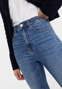 Jeans de mezclilla azul de cintura alta con una textura suave y bolsillos delanteros, con costuras doradas y un cierre de botón plateado.