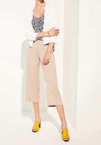 Chaqueta blanca sobre una blusa de estampado en blanco y negro, combinada con pantalones anchos beige y mules amarillos de punta abierta con detalles en negro.