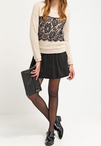 Pull beige avec superposition en dentelle noire, jupe évasée noire, collants noirs et bottines brillantes noires. Tenant une pochette noire.