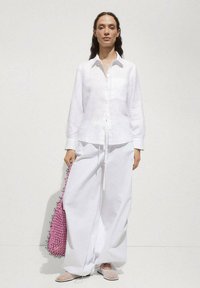 Cămașă albă din in cu nasturi, asociată cu pantaloni albi lejeri. Accesorizată cu o geantă împletită roz. Textură netedă și croială lejeră.