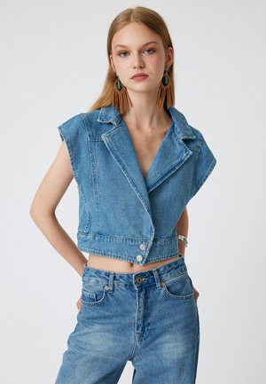 Chaleco de denim recortado en azul claro, con mangas cortas, cuello con muescas y dos botones delanteros. Combinado con unos pantalones vaqueros de cintura alta a juego.