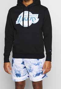 Czarna bluza z kapturem z logo Los Angeles Lakers w jasnoniebieskim kolorze, z kieszenią na froncie i sznurkiem do regulacji. W zestawieniu z białymi szortami w wzór chmur.