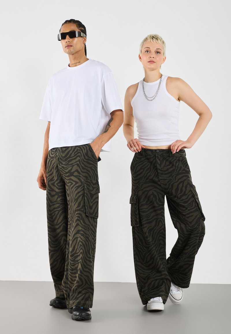 Cargo pants in green with black zebra stripes, paired with a white oversized t-shirt and a white tank top. Sunglasses and silver chain accents.