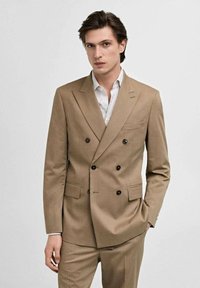 Completo beige doppiopetto con rever a lancia, due tasche frontali e bottoni neri. Abbinato a una camicia bianca a righe azzurro chiaro.