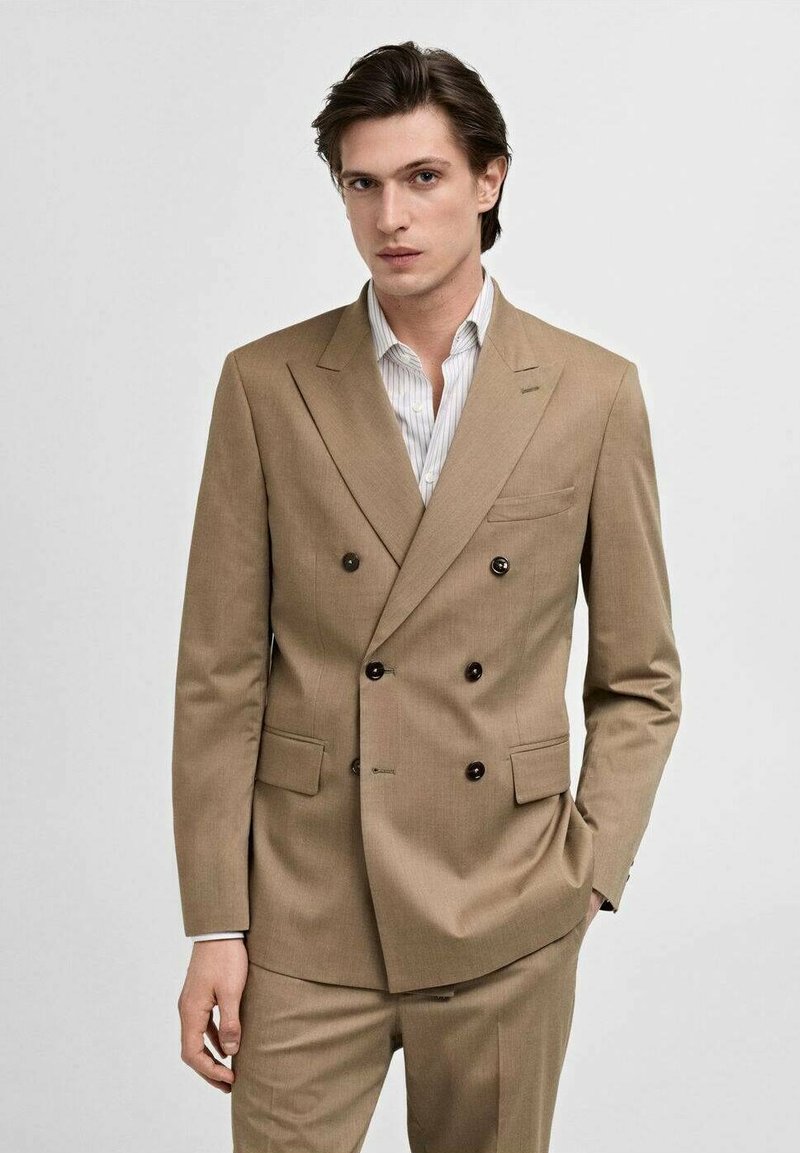 Completo beige doppiopetto con rever a lancia, due tasche frontali e bottoni neri. Abbinato a una camicia bianca a righe azzurro chiaro.