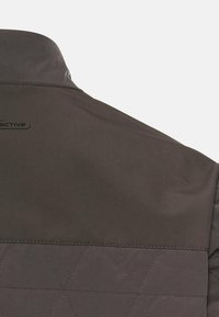 Grå overtøj med en quiltet tekstur på den nederste del, glat stof på den øverste del og en kort krave med branding.
