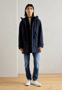 Navy blue parka with a hood, front zipper, drawstring waist, and two side pockets. Worn over a light top and paired with blue jeans and white sneakers.