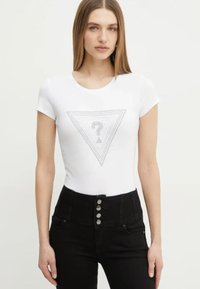 T-shirt blanc à manches courtes arborant un design graphique triangulaire avec un point d'interrogation au centre, associé à un jean noir taille haute.