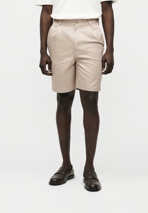 Άνδρας που φοράει μπεζ σορτς ραμμένα στα μέτρα του, λευκό κοντομάνικο πουκάμισο και σκούρα καφέ δερμάτινα μοκασίνια, στέκεται μπροστά σε μονόχρωμο φόντο.