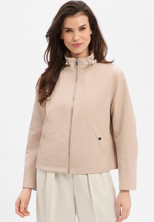 Kobieta ubrana w beżową kurtkę na zamek błyskawiczny z zapinanymi na guziki bocznymi kieszeniami i wysokim kołnierzem, zestawiona z jasnymi, szerokimi spodniami, stojąca na białym tle.