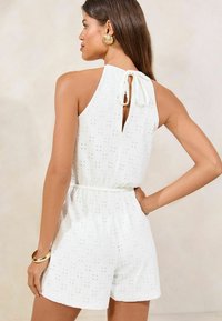 Romper sans manches blanc avec motif ajouré, détail à nouer dans le dos, taille élastique et shorts amples. Il présente une texture lisse et un design simple.