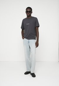 Grijze T-shirt met korte mouwen en witte tekst, lichtblauwe recht-toe-rechtdoor jeans en zwarte schoenen. Model draagt donkere zonnebril en staat met de handen in de zakken.