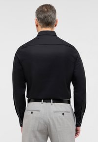 Schwarzes Langarmhemd mit geradem Kragen, glatter Textur und geknöpften Manschetten, kombiniert mit hellgrauen Hosen mit Hintertaschen.