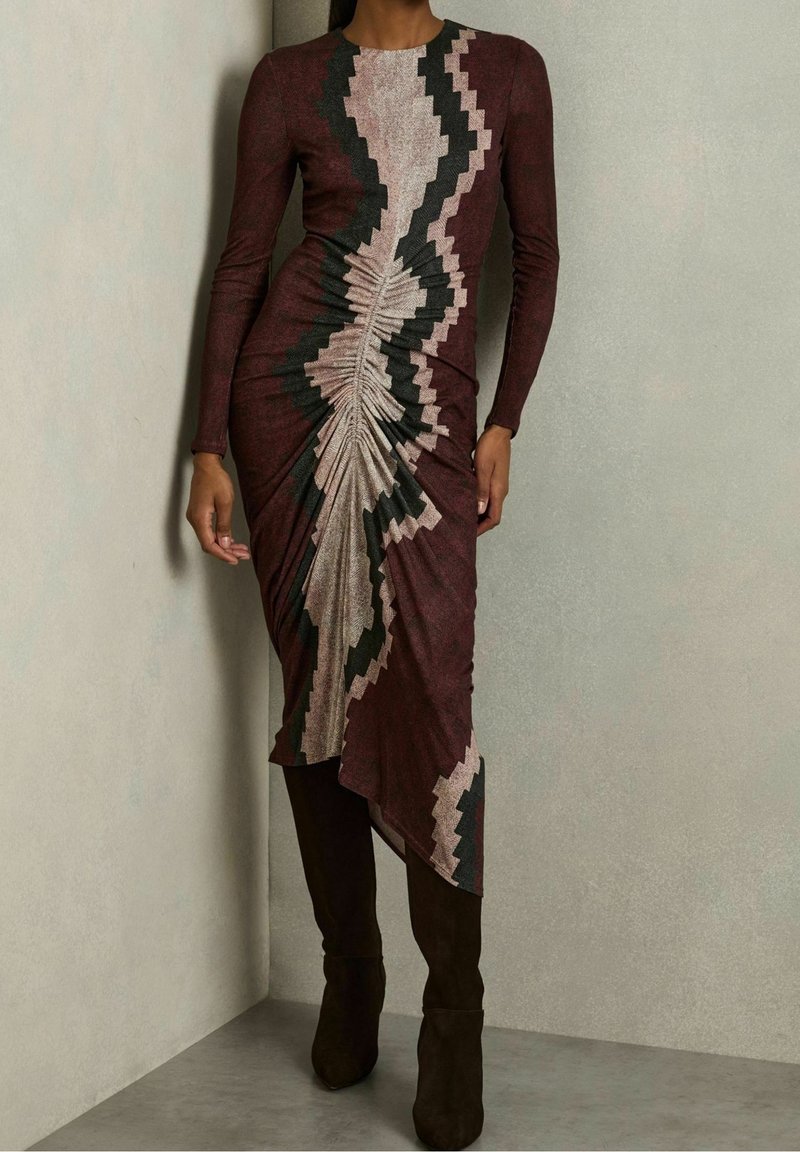 Robe à manches longues et ajustée, présentant un motif en zigzag aux nuances de bordeaux, noir et rose, avec une texture froncée et un ourlet asymétrique.