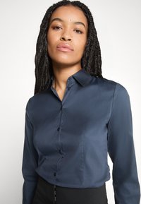Camicia blu navy con bottoni realizzata in tessuto liscio, con maniche lunghe, colletto a punta e vestibilità slim. Presenta una tasca e bottoni discreti.