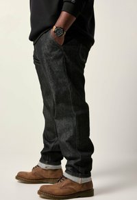 Uomo che indossa jeans scuri arrotolati, scarpe di pelle marroni, una camicia nera a maniche lunghe, con le mani nelle tasche e un orologio al polso sinistro.