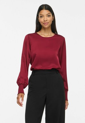 Femme aux longs cheveux foncés, portant un chemisier bordeaux à manches longues et un pantalon noir taille haute, debout sur un fond blanc.