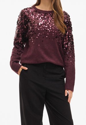 Kvinde iført en mørkelilla sweater med lyserøde pailletter på skuldre og øvre ærmer, kombineret med sorte bukser med høj talje.
