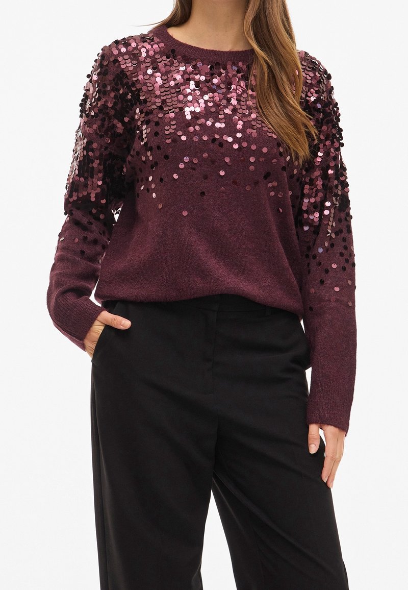 Femme portant un pull violet foncé avec des sequins roses aux épaules et sur le haut des manches, associé à un pantalon noir taille haute.