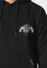 Felpa nera con tasca frontale e coulisse, con testo "Pas de Monaco", bandiere a scacchi e grafica di una corona di alloro sul petto.