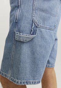 Denim cargo shorts in light blue with a straight shape, side pockets, and rolled hem. Features visible stitching and texture details.
