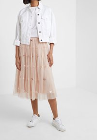 Een vrouw draagt een witte cropped jacket over een witte shirt, een beige tulle midi rok met pailletten, en witte sneakers, staand in een minimalistische omgeving.