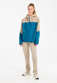 Ľahká vonkajšia bunda v teal a béžovej farbe, s predným zipsom, nastaviteľnou kapucňou a elastickými manžetami. Kombinované s béžovými nohavicami a teniskami.