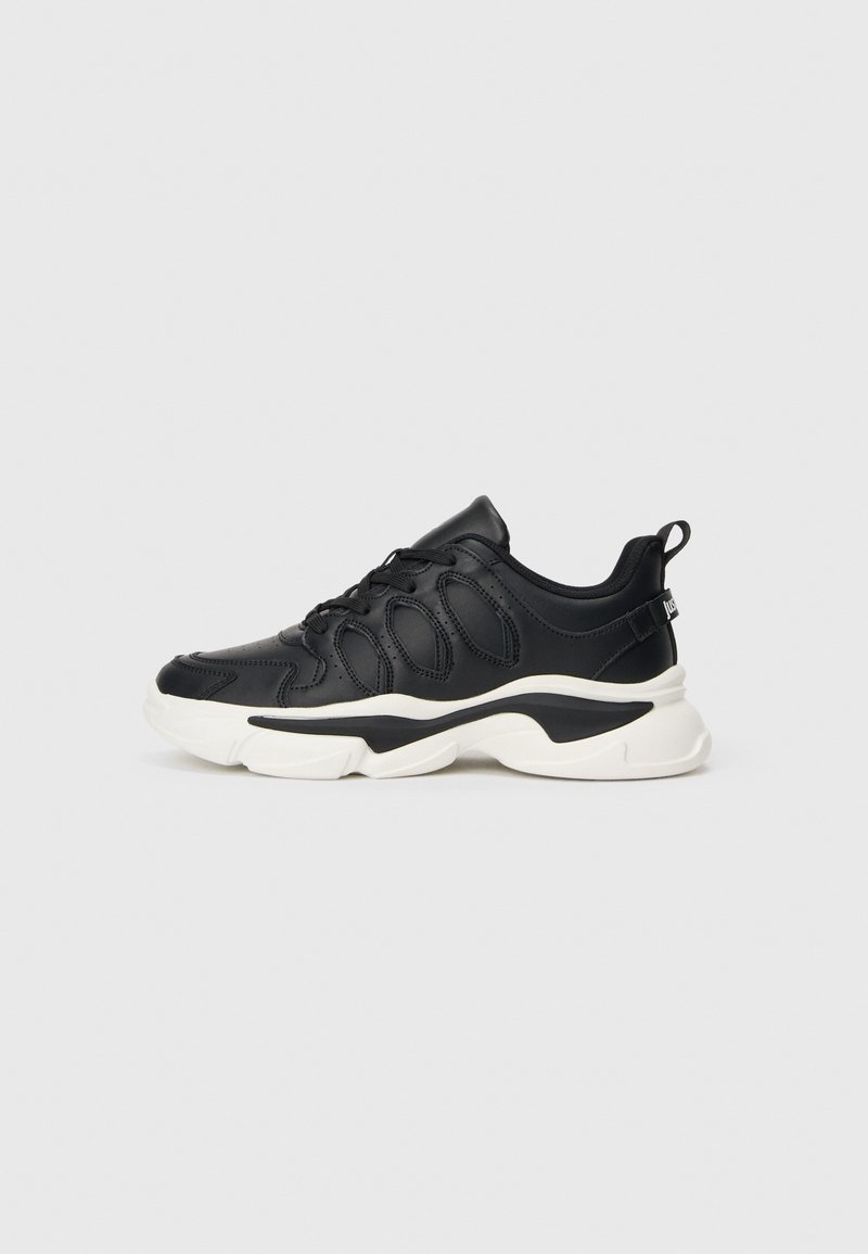 Svart lavtoppsneaker med hvit kraftig såle, snøring foran og en løkke på hælen, vist mot en enkel lys bakgrunn.
