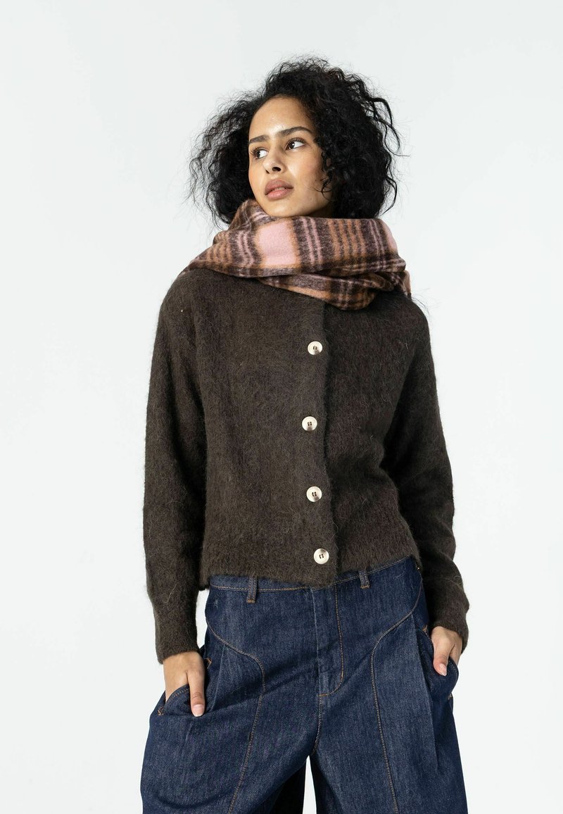 Femme aux cheveux bouclés portant un pull marron boutonné, une écharpe à carreaux rose et marron, et un jean bleu ample, les mains dans les poches.