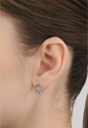 Primer plano de una oreja con piel lisa y cabello castaño, que lleva un pendiente circular de plata incrustado con pequeñas piedras preciosas transparentes.