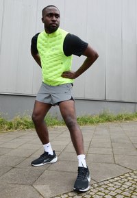 Hombre con chaleco verde brillante y pantalones cortos grises estirando las piernas al aire libre sobre el pavimento cerca de la pared de un edificio.