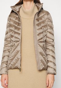 Frau in einem beigen, gerippten Strick-Turtleneck-Kleid und einer hellbraunen, gesteppte Daunenjacke mit Reißverschlusstaschen, vor einem weißen Hintergrund stehend.