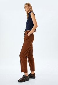 Dunkelblauses ärmelloses Oberteil, braune hochgewaistete Hose und braune Schnürschuhe. Die Hose hat eine gerade Passform und seitliche Taschen.