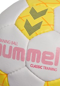 Vit och gul träningsfotboll med en texturerad yta, djärv rosa logotyp och ett chevronmönster i blått och svart.