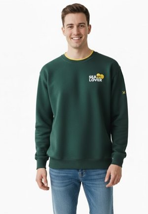 Joven sonriente que lleva una sudadera verde oscuro con el texto "SEA LOVER" y detalles amarillos, combinado con vaqueros azules, posando frente a un fondo blanco.