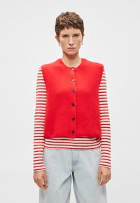 Κόκκινο πλεκτό γιλέκο με ραβδωτή υφή, στρογγυλή λαιμόκοψη και κουμπιά μπροστά. Φοριέται πάνω από ένα μακρυμάνικο πουκάμισο με ρίγες σε λευκό και κόκκινο χρώμα.