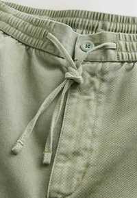 Ceinture de pantalon vert clair avec bande élastique, fermeture par bouton et cordon noué en nœud.