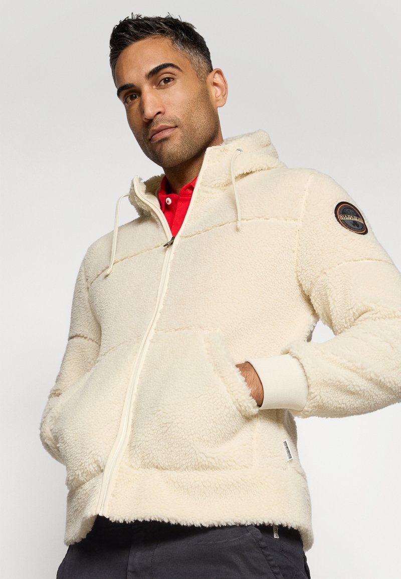 Mann trägt eine cremefarbene Fleecejacke mit Reißverschluss und Kapuze, die Hände in den Taschen, und ein rotes Hemd darunter vor einem schlichten Hintergrund.