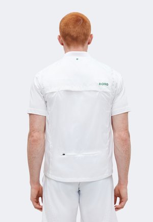 Muž so zrzavými vlasmi otočený chrbtom, nosiaci biele tričko s krátkym rukávom s logom "BORG" na hornej časti chrbta a biele šortky.