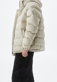 Beige pufferjack met capuchon, voorzien van doorgestikte delen, elastische manchetten en een logopatch op de mouw. Gecombineerd met zwarte cargobroek.