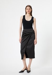 Top nero senza maniche abbinato a una gonna di satin scura all'altezza del ginocchio con un dettaglio attorcigliato in vita e uno spacco frontale. Ballerine nere appuntite.