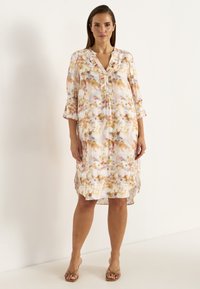 Blumenmusterkleid aus weichem Stoff, mit V-Ausschnitt, Rüschendetails, Knopfverschluss und Dreiviertelärmeln. Helle Pastellfarben dominieren.